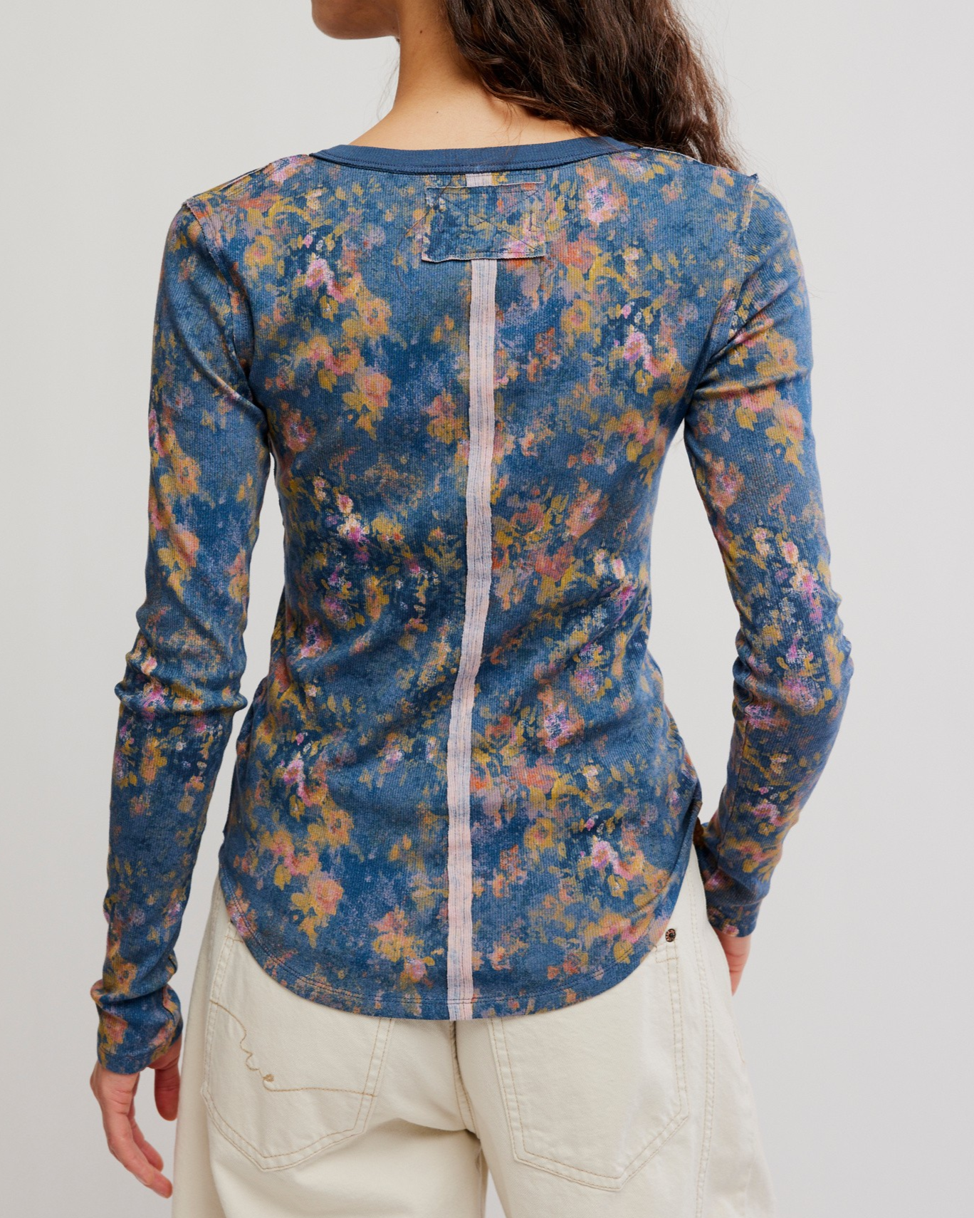 A woman shown from the back wearing a long-sleeve floral top with a blue base and multicolor floral print, paired with light beige pants, against a plain light gray background.