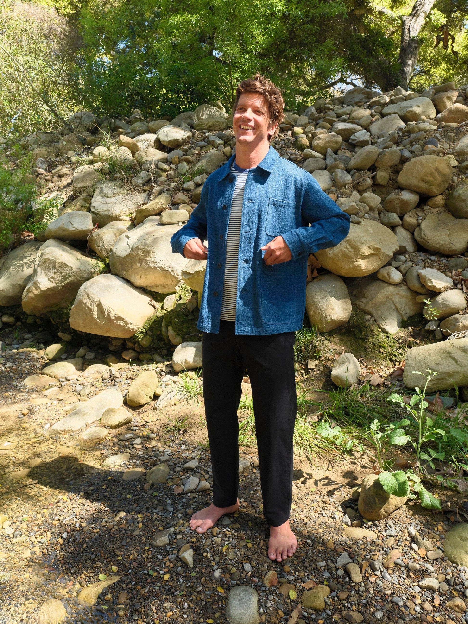 Person standing outdoors in front of a stone wall with greenery