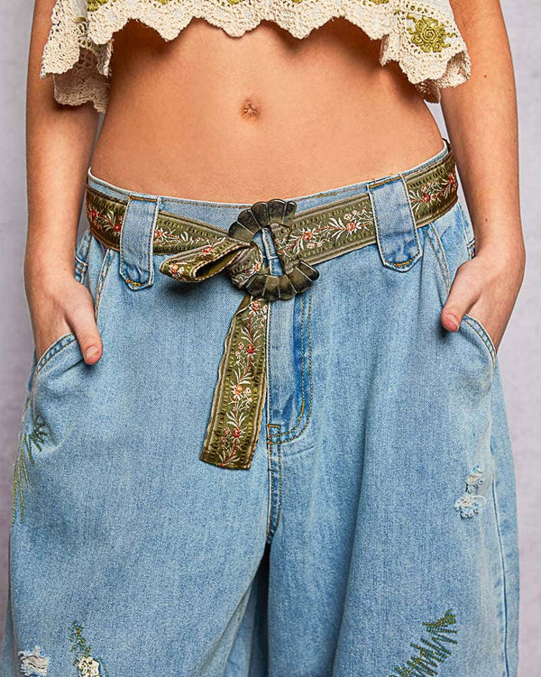Person wearing a floral embroidered belt with a white top and blue jeans on a light gray background