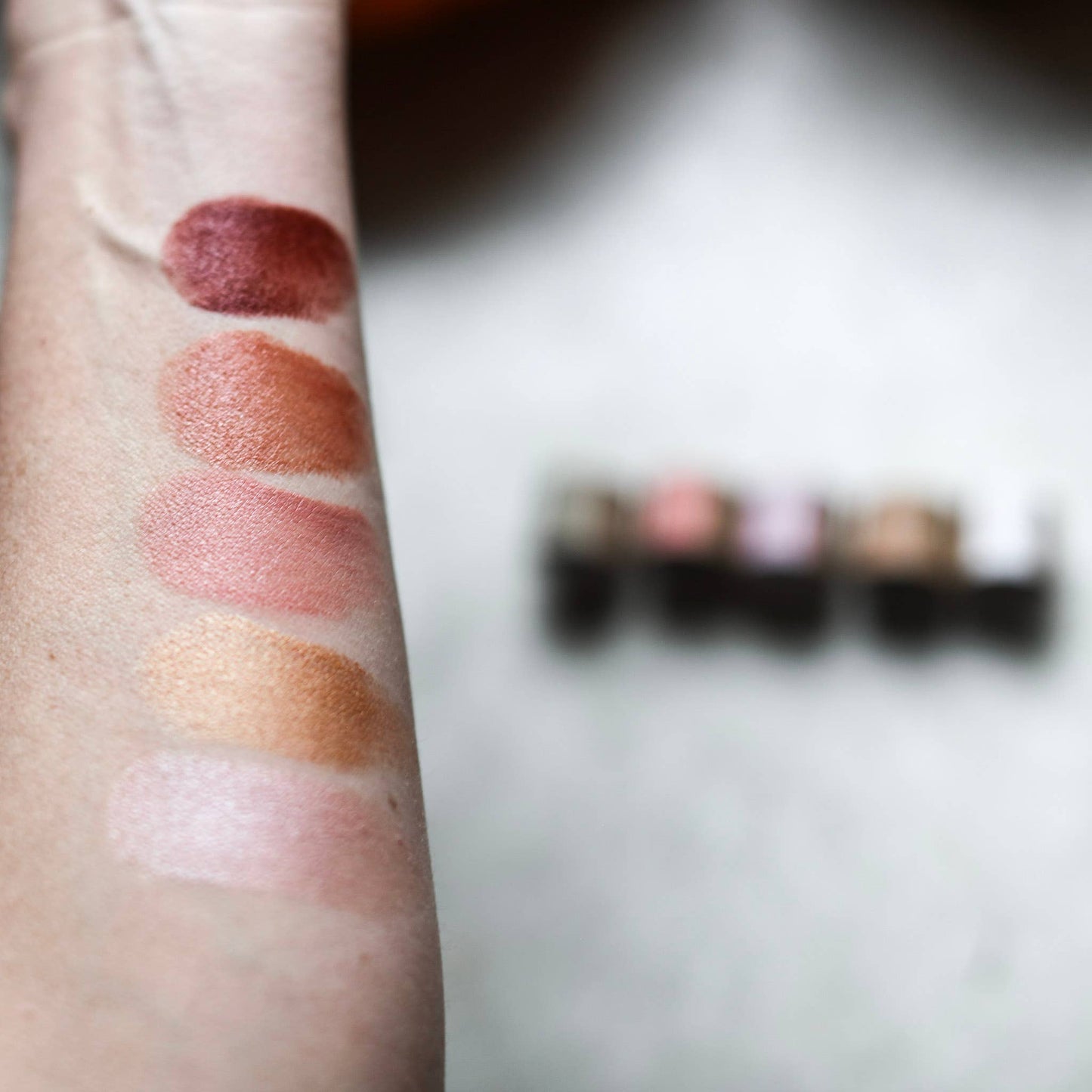Lipstick swatches on a person's arm with a blurred makeup palette in the background