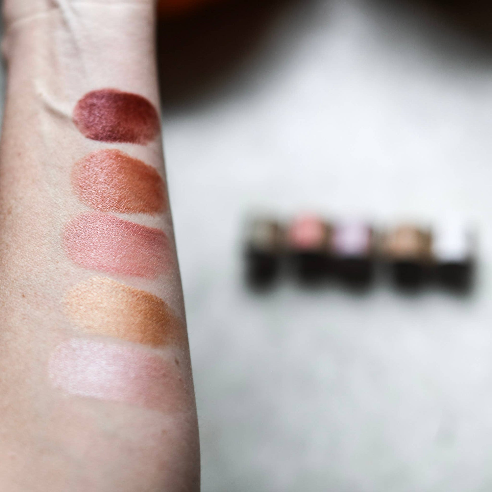 Lipstick swatches on a person's arm with a blurred makeup palette in the background