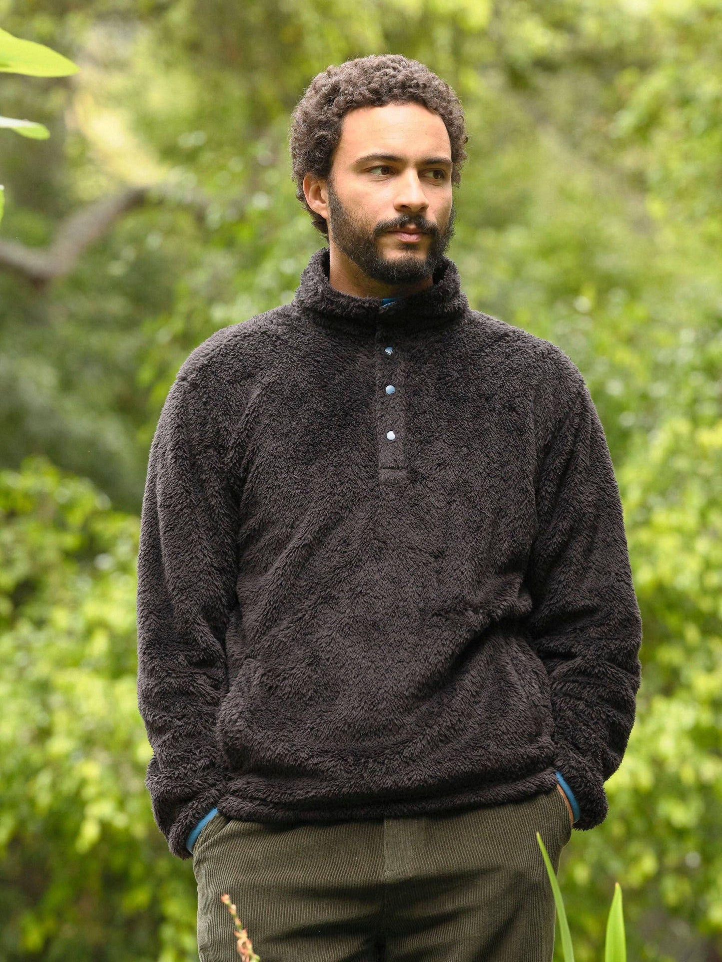 Man wearing a dark fleece pullover standing in a natural setting with greenery.