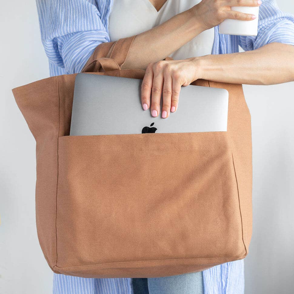 A person wearing a blue and white striped shirt holds a large tan canvas tote bag with a laptop tucked into the front pocket. The person is also holding a white cup.