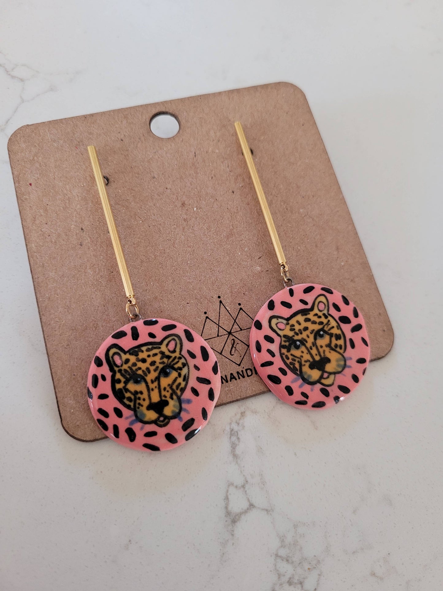 Pair of earrings with leopard design on pink background, displayed on a cardboard card.