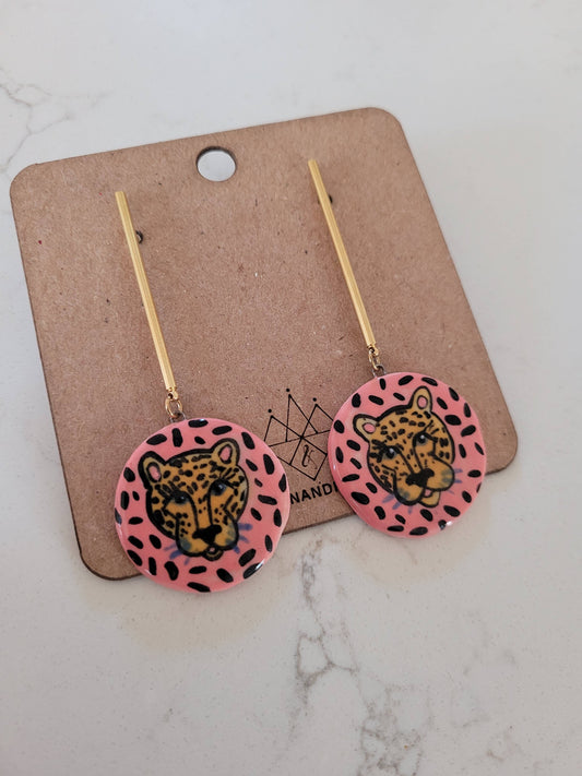 Pair of earrings with leopard design on pink background, displayed on a cardboard card.