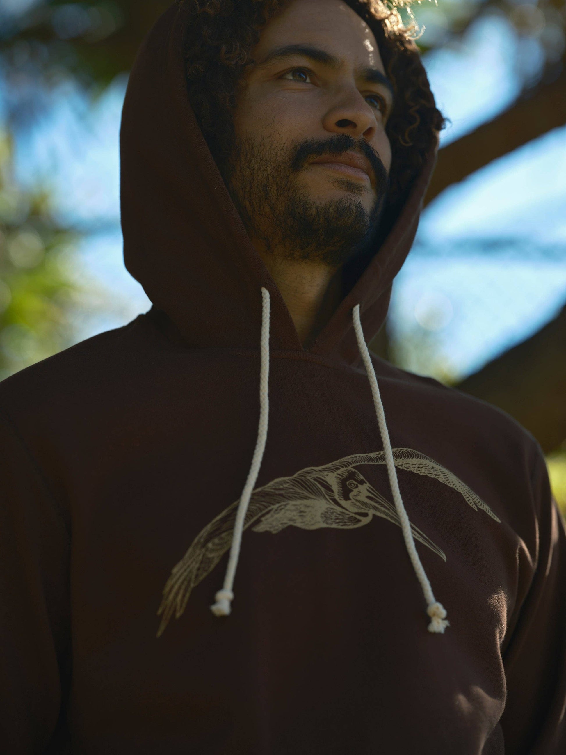 Man wearing a brown hoodie with a logo, standing outdoors with blurred background