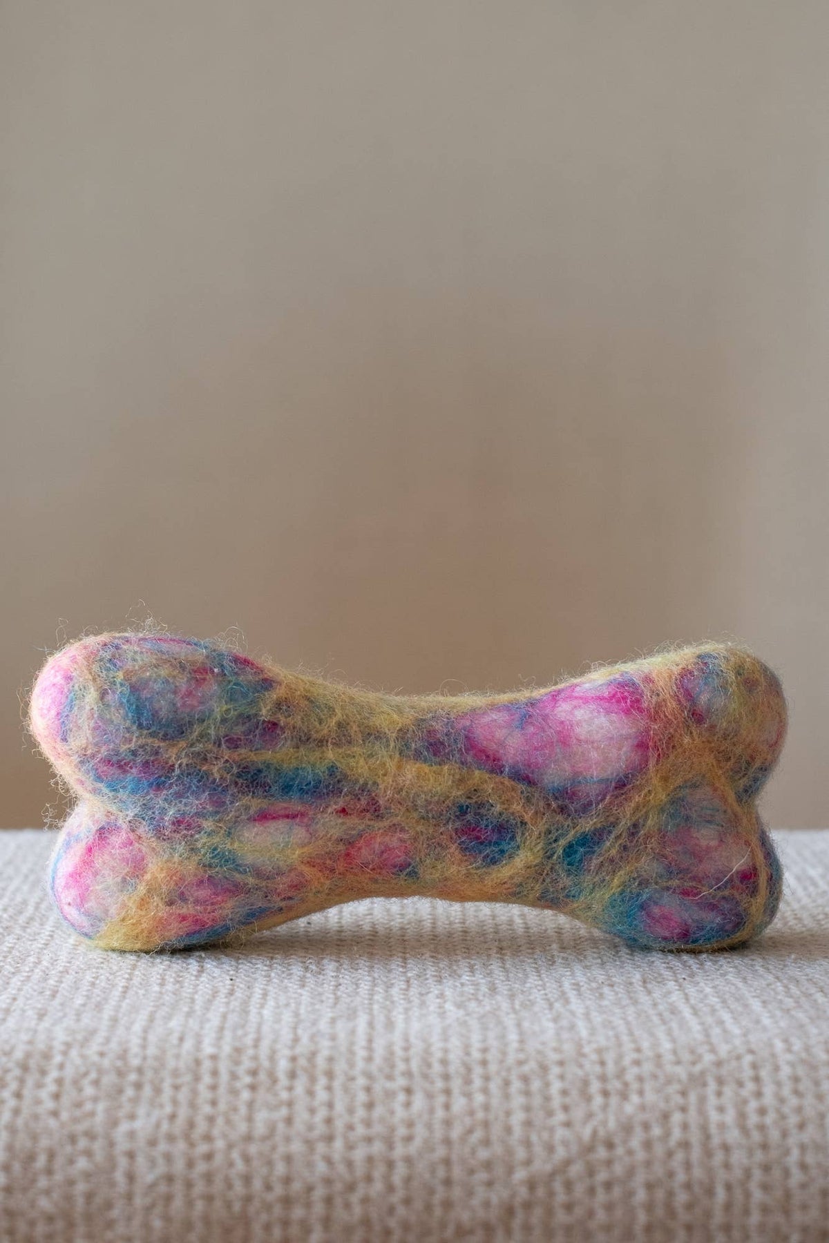 A handmade bone-shaped felted wool pet toy with a marbled pattern of pink, blue, yellow, and white fibers, resting on a textured beige fabric surface against a neutral beige background.