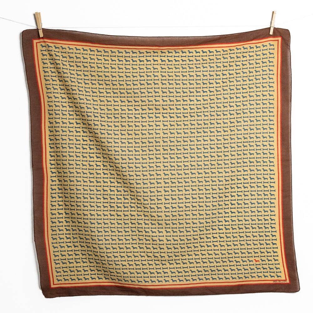 A square scarf with a repeating geometric pattern in beige, brown, and orange, finished with a solid dark brown border, hanging from two wooden clothespins against a white background.