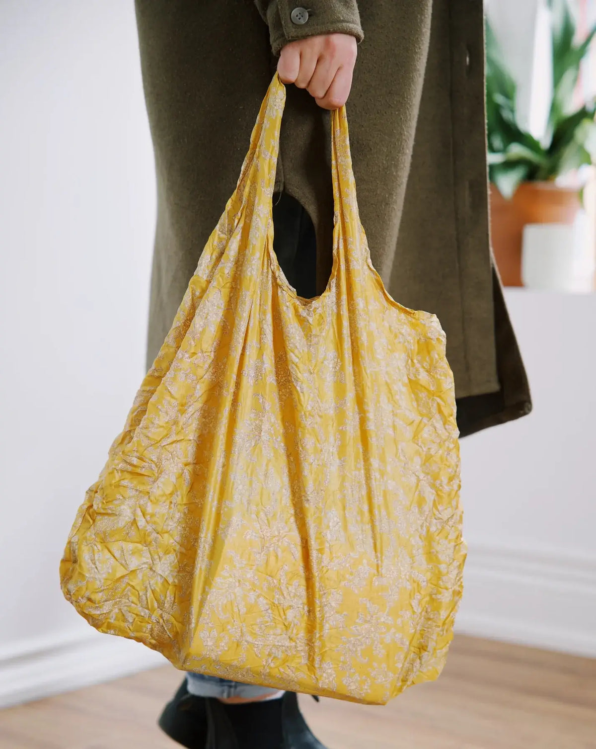A person holding a large mustard yellow reusable tote bag with a white floral pattern, wearing an olive green coat and black boots in a bright indoor setting.