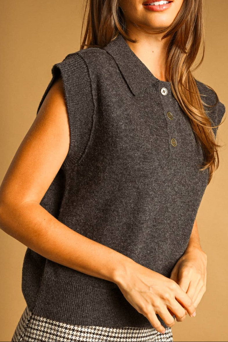 A woman wearing a dark gray sleeveless knit polo vest, posing against a solid warm brown studio background.