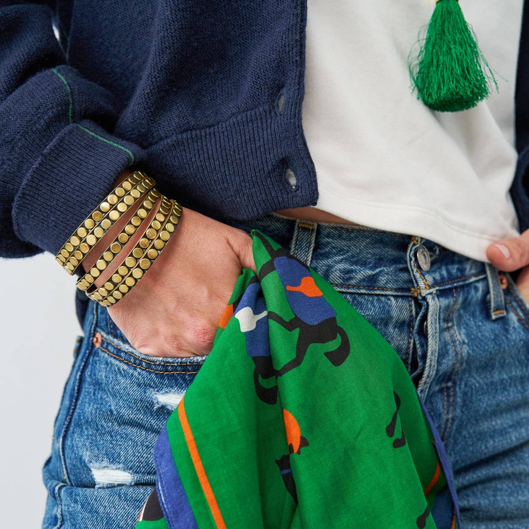 A close-up lifestyle shot of a person wearing a navy blue cardigan, white t-shirt, and distressed blue jeans, accessorized with gold beaded bracelets and holding a green patterned scarf.