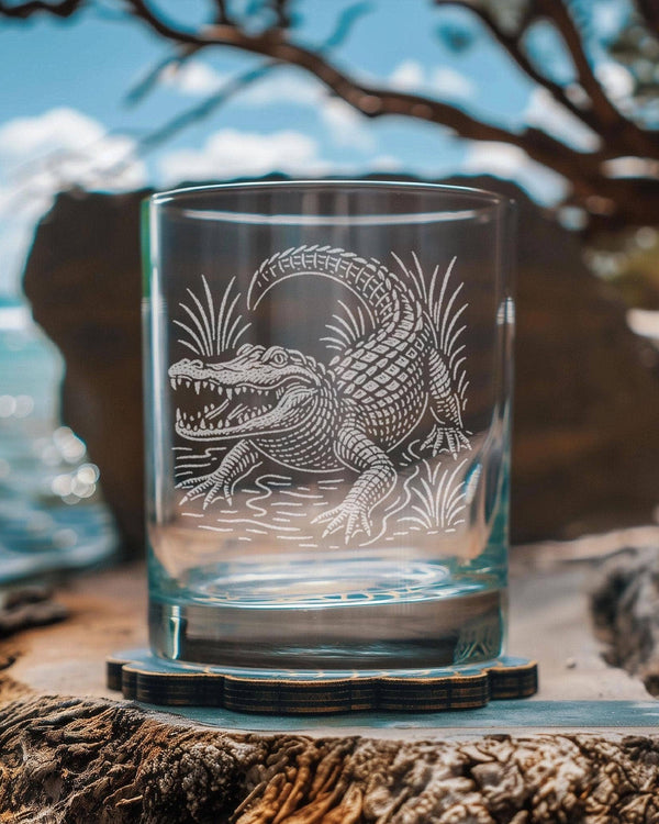 A clear glass rocks tumbler with an etched alligator design, resting on a wooden slice coaster outdoors on a beach with rocks and ocean in the background.