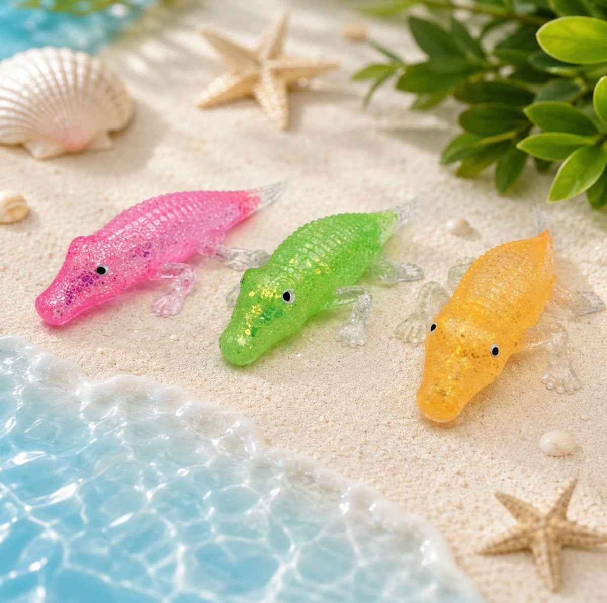 Three translucent alligator toys in pink, green, and yellow are arranged on a sandy beach scene with starfish, seashells, and blue water.