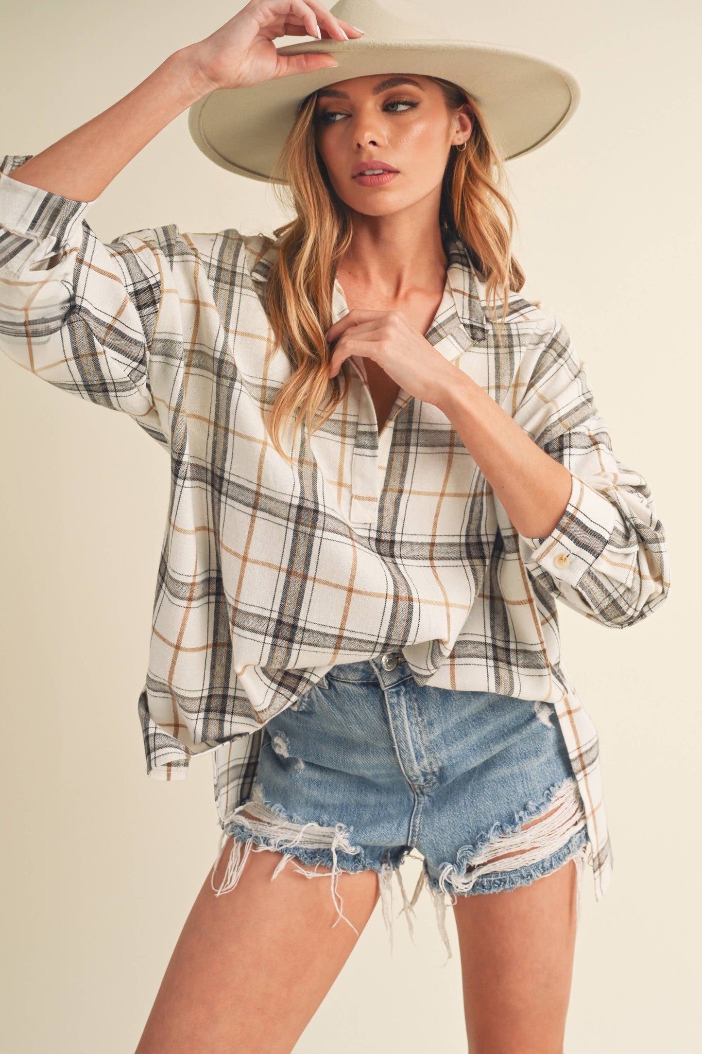 Woman wearing a plaid shirt, denim shorts, and a wide-brimmed hat on a plain background