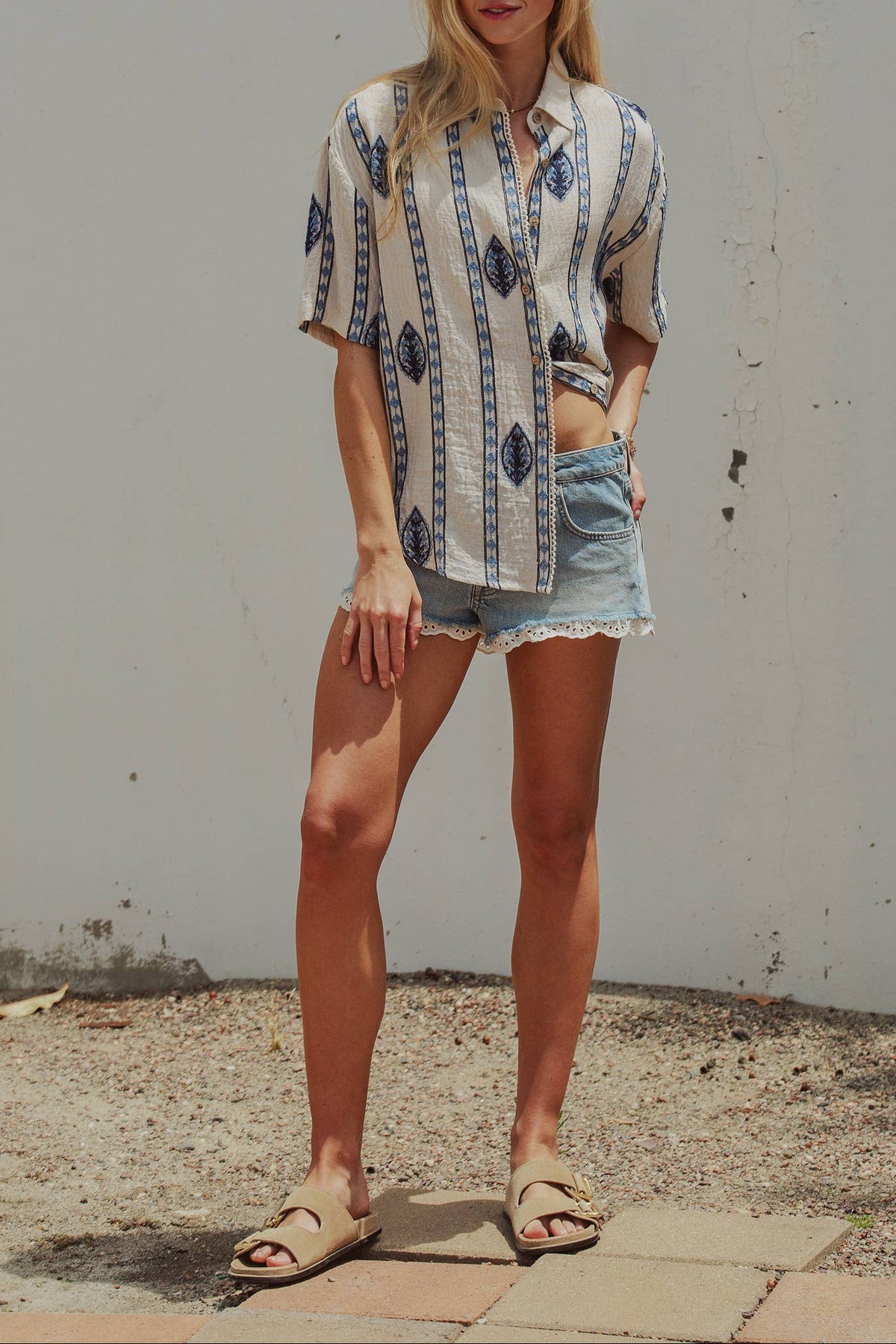 Woman wearing a patterned shirt and denim shorts standing against a plain wall.