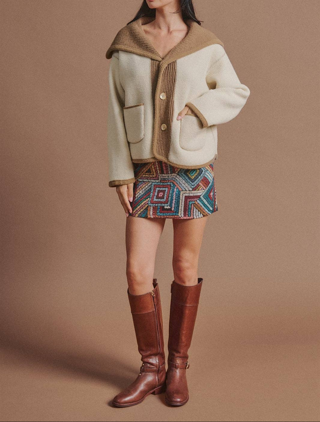 A model stands against a solid brown studio backdrop wearing a cream and brown textured jacket with a brown cap, a colorful geometric patterned mini skirt, and brown leather knee-high boots.