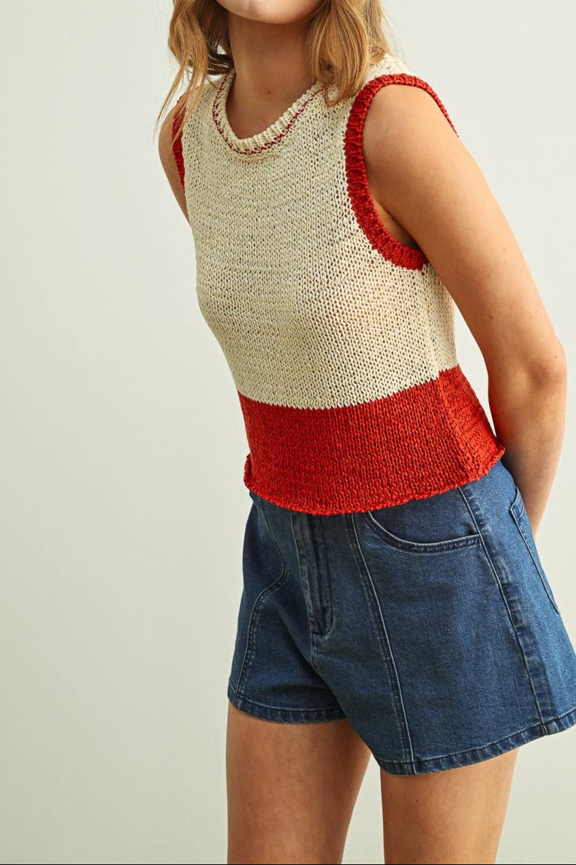 Woman wearing a sleeveless, color-blocked top with beige and red sections, paired with denim shorts on a plain background.