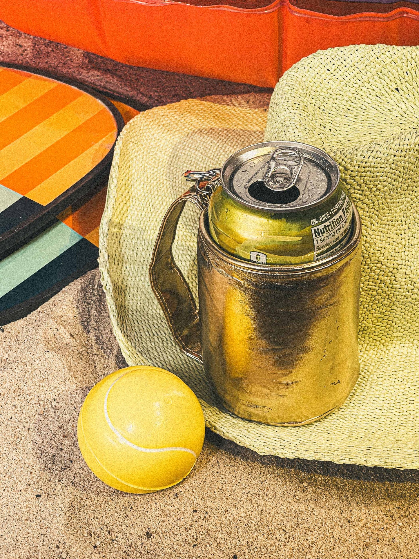 Gold thermos with a can on a straw hat next to a yellow tennis ball on sand