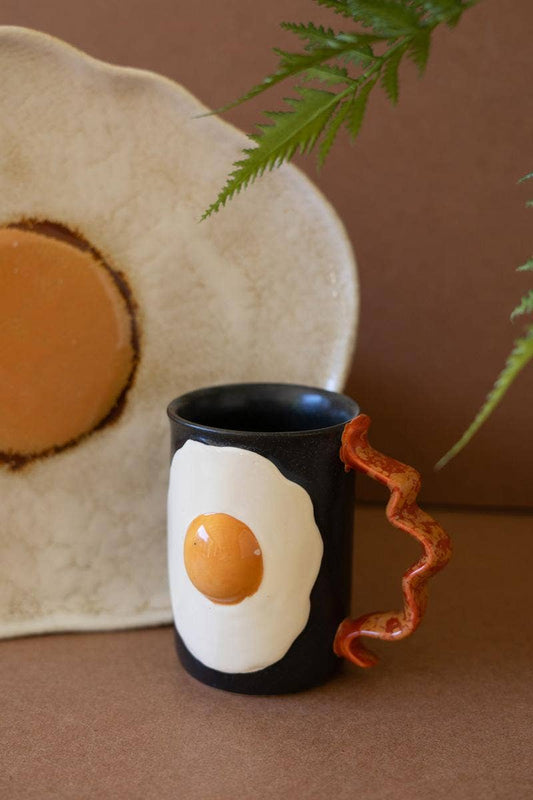 Black mug with a design of an egg and bacon next to a white plate with a brown circle on a brown background