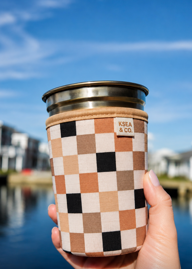 A hand holding a stainless steel tumbler with a beige and black checkerboard patterned fabric sleeve. The sleeve features a small tan leather patch with the text 'KSEA & CO.