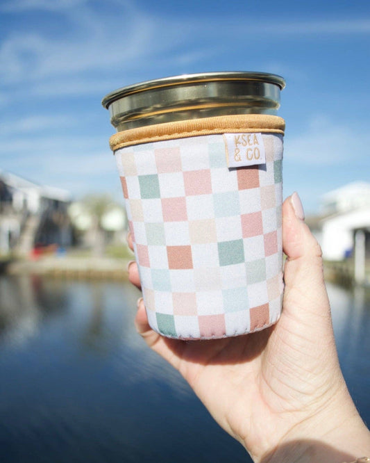A hand holding a reusable glass can cooler with a pastel checkerboard pattern, a tan leather-like sleeve, and a clear lid, set against a blurred outdoor background of water and buildings.