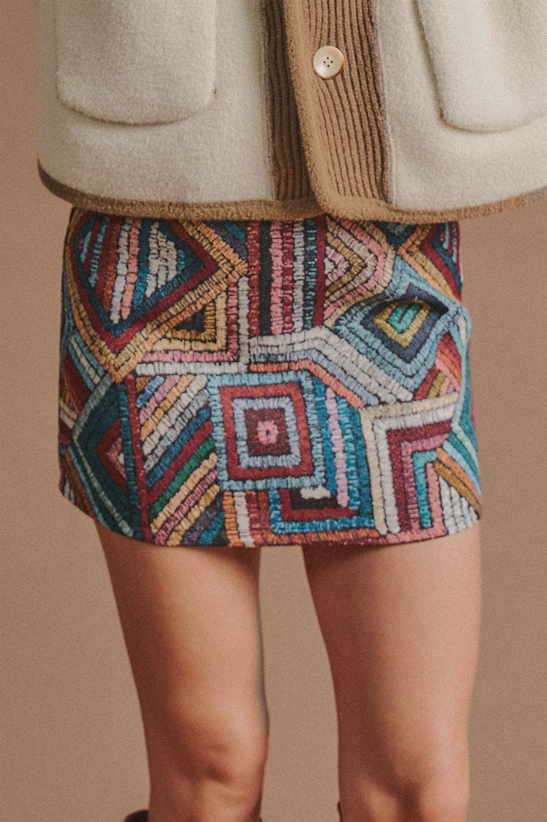 A close-up of a person wearing a short, highly textured geometric patterned skirt in red, blue, beige, and green, paired with a cream-colored jacket.