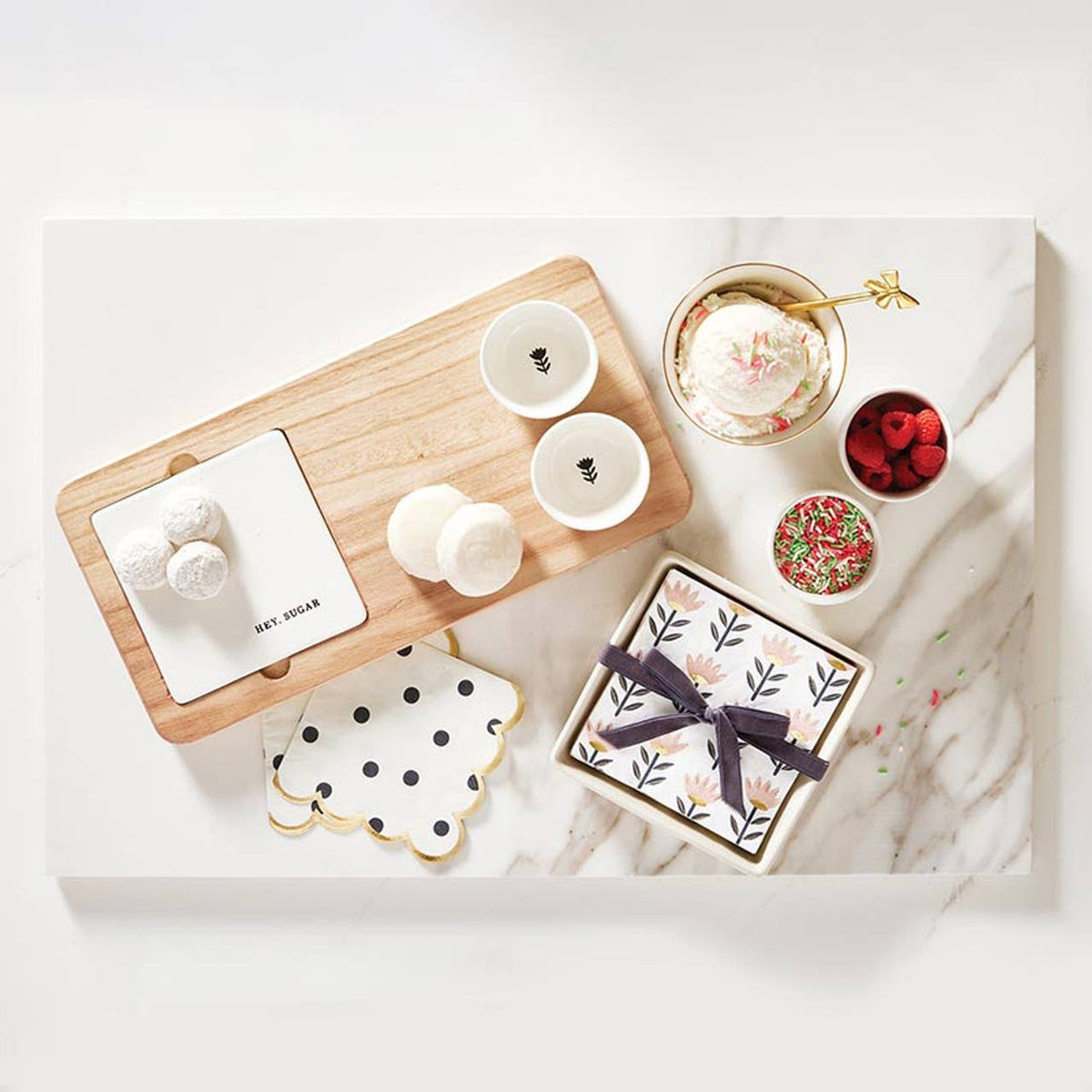An overhead flat-lay of assorted sweet treats and gift items arranged on a white marble board. The set includes a wooden serving tray with white sugar-coated pastries, three small white cups, a bowl of ice cream with whipped cream and pink sprinkles, a bowl of red raspberries, a small bowl of colorful sprinkles, a patterned gift box tied with a dark ribbon, and a scalloped napkin with black polka dots.