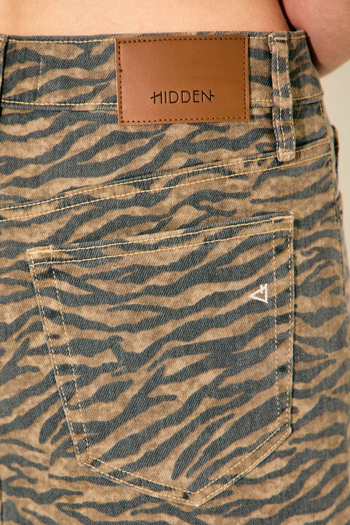 Close-up of the back waistband and pocket area of patterned denim jeans, showing a tan leather patch with the brand name HIDDEN and a small white geometric logo on the pocket.