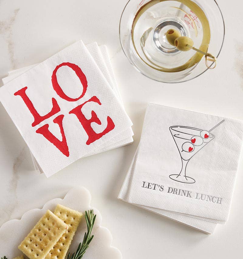 Two white paper napkins styled on a light marble surface. One napkin has the word LOVE printed in large red letters, and the other has a martini glass illustration with olives and the text LET'S DRINK LUNCH.