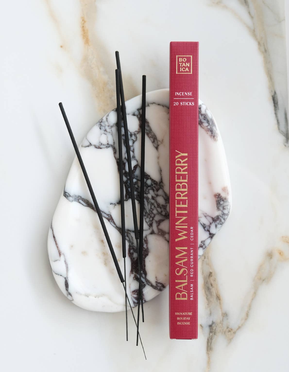 A red rectangular box of Spitz & Tanca Balsam Winterberry incense sticks sits on a white marble tray with black incense sticks, arranged on a white marble surface.