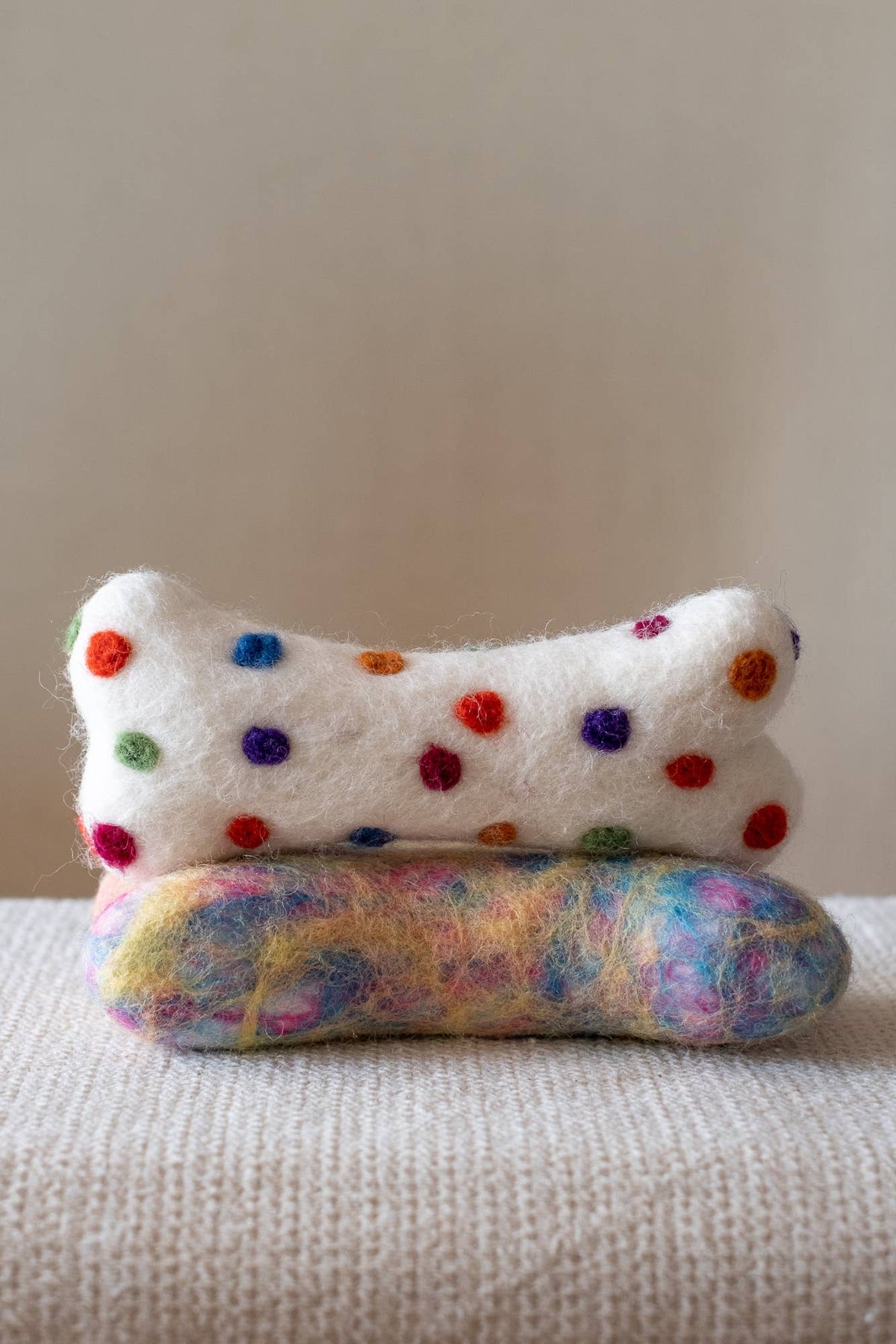 Two handmade felted wool dog toys shaped like bones, stacked on a textured white surface against a neutral beige background. The top toy is white with colorful felted polka dots in red, orange, blue, green, and purple.