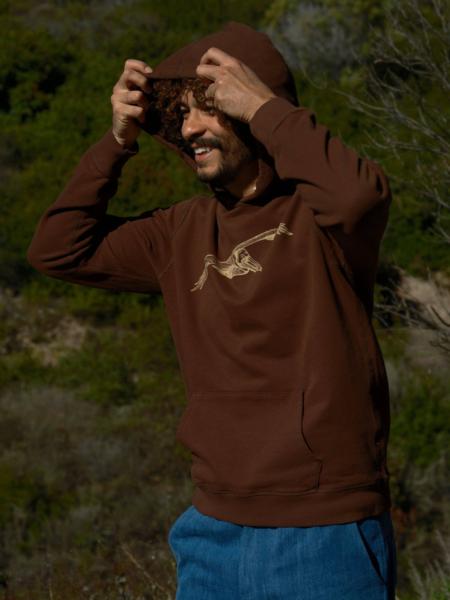 Person wearing a brown hoodie with a logo, standing outdoors in a natural setting.