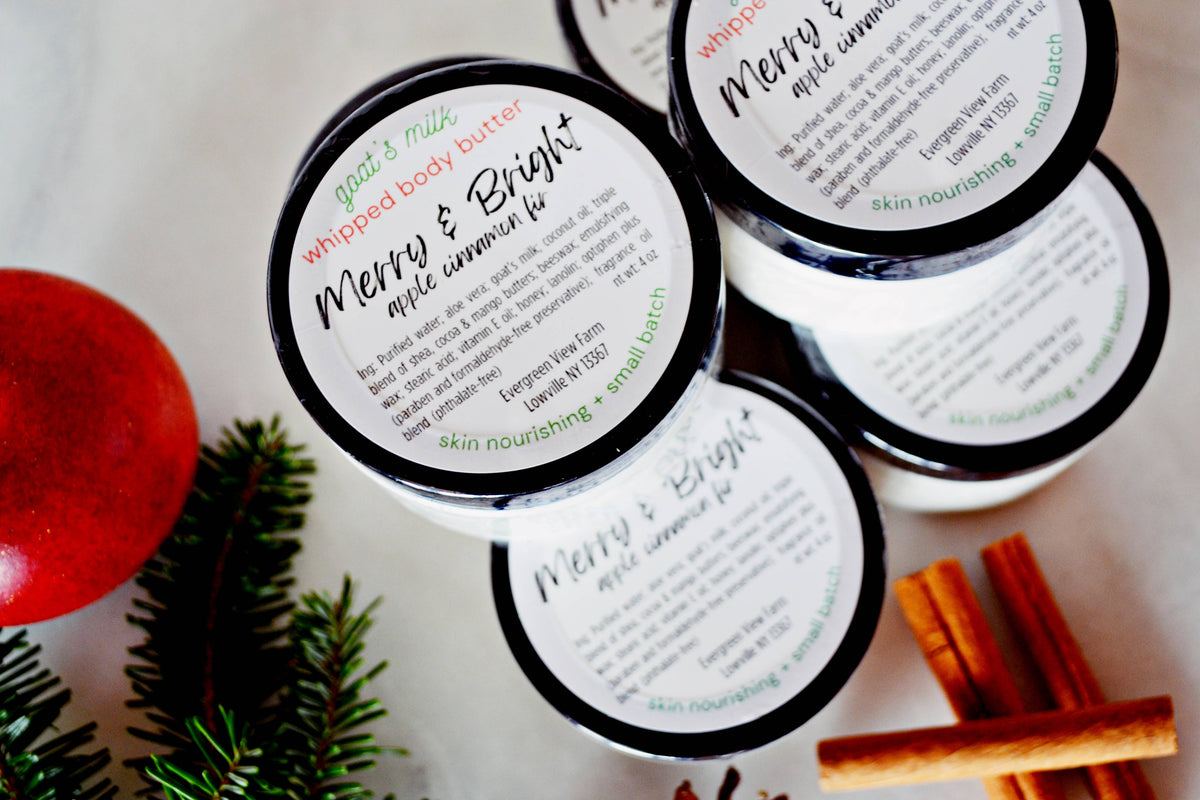 Four jars of God's Mix Whipped Body Butter arranged on a light surface with holiday-themed props including a red ornament, pine branches, and cinnamon sticks. The lids feature white labels with black and green text, highlighting the 'Merry & Bright' apple cinnamon scent.