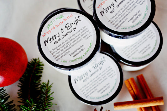 Four jars of God's Mix Whipped Body Butter arranged on a light surface with holiday-themed props including a red ornament, pine branches, and cinnamon sticks. The lids feature white labels with black and green text, highlighting the 'Merry & Bright' apple cinnamon scent.