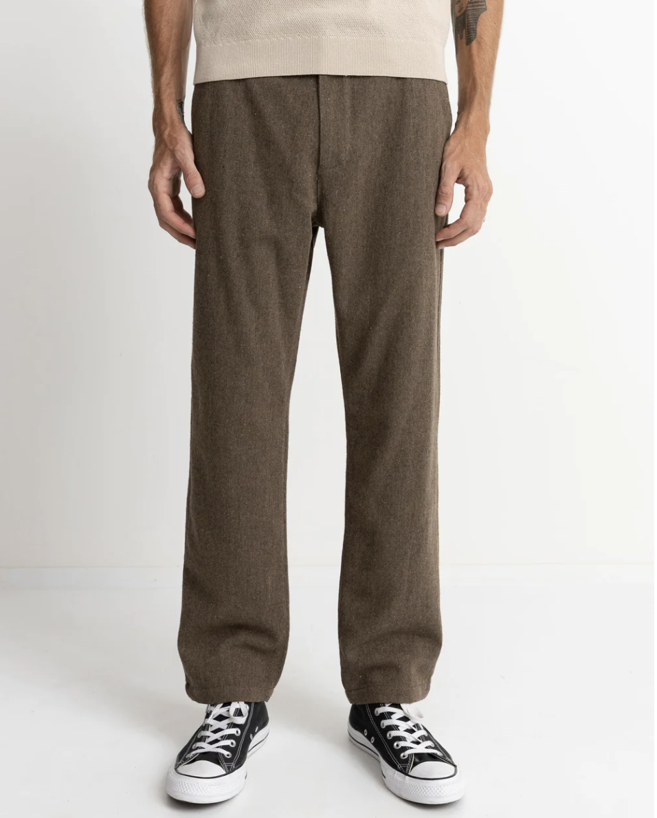 A person standing and wearing the Essential Trouser in brown with a fixed waist and straight leg fit, paired with a beige shirt and black and white sneakers.