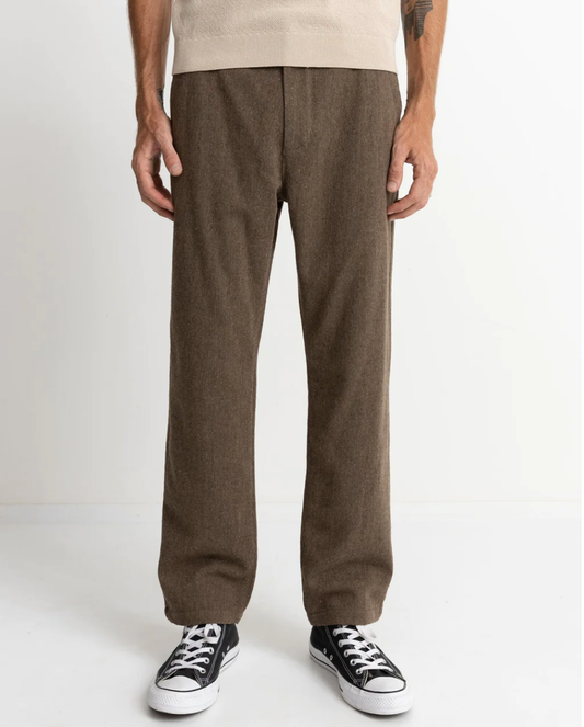 A person standing and wearing the Essential Trouser in brown with a fixed waist and straight leg fit, paired with a beige shirt and black and white sneakers.