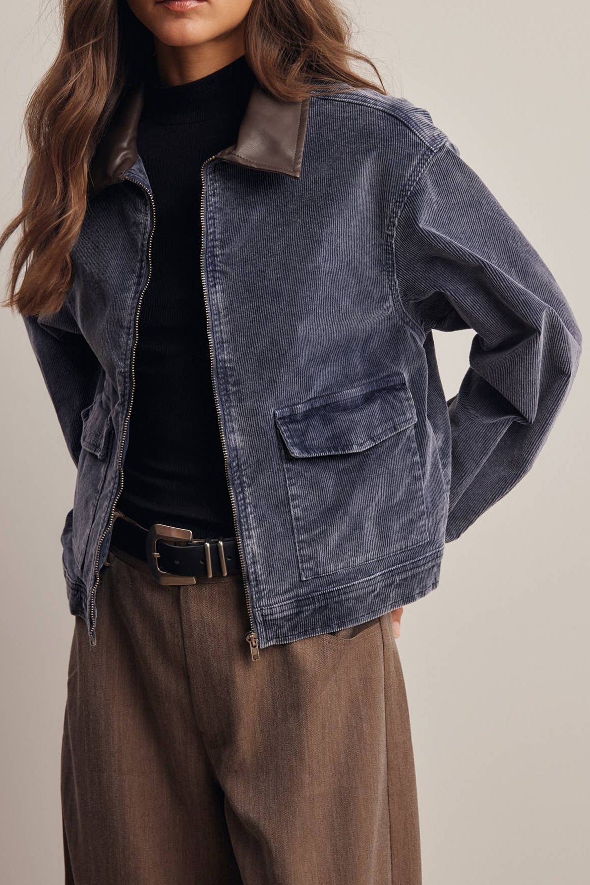 A woman modeling a washed blue denim jacket with brown leather shoulder tabs, worn over a black top and brown trousers, against a solid beige studio background.