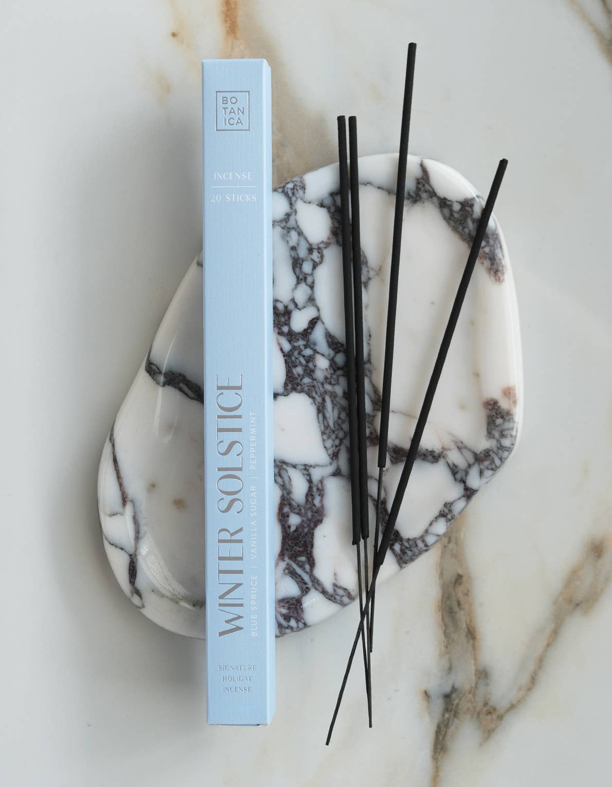 A light blue rectangular box labeled 'WINTER SOLSTICE' stands upright on a white marble tray with black veining, with several thin black reeds placed on top, all on a white marble surface.