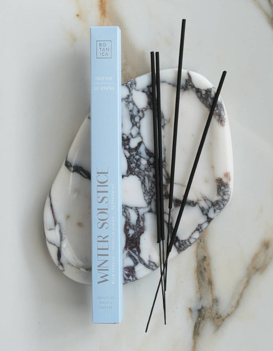 A light blue rectangular box labeled 'WINTER SOLSTICE' stands upright on a white marble tray with black veining, with several thin black reeds placed on top, all on a white marble surface.