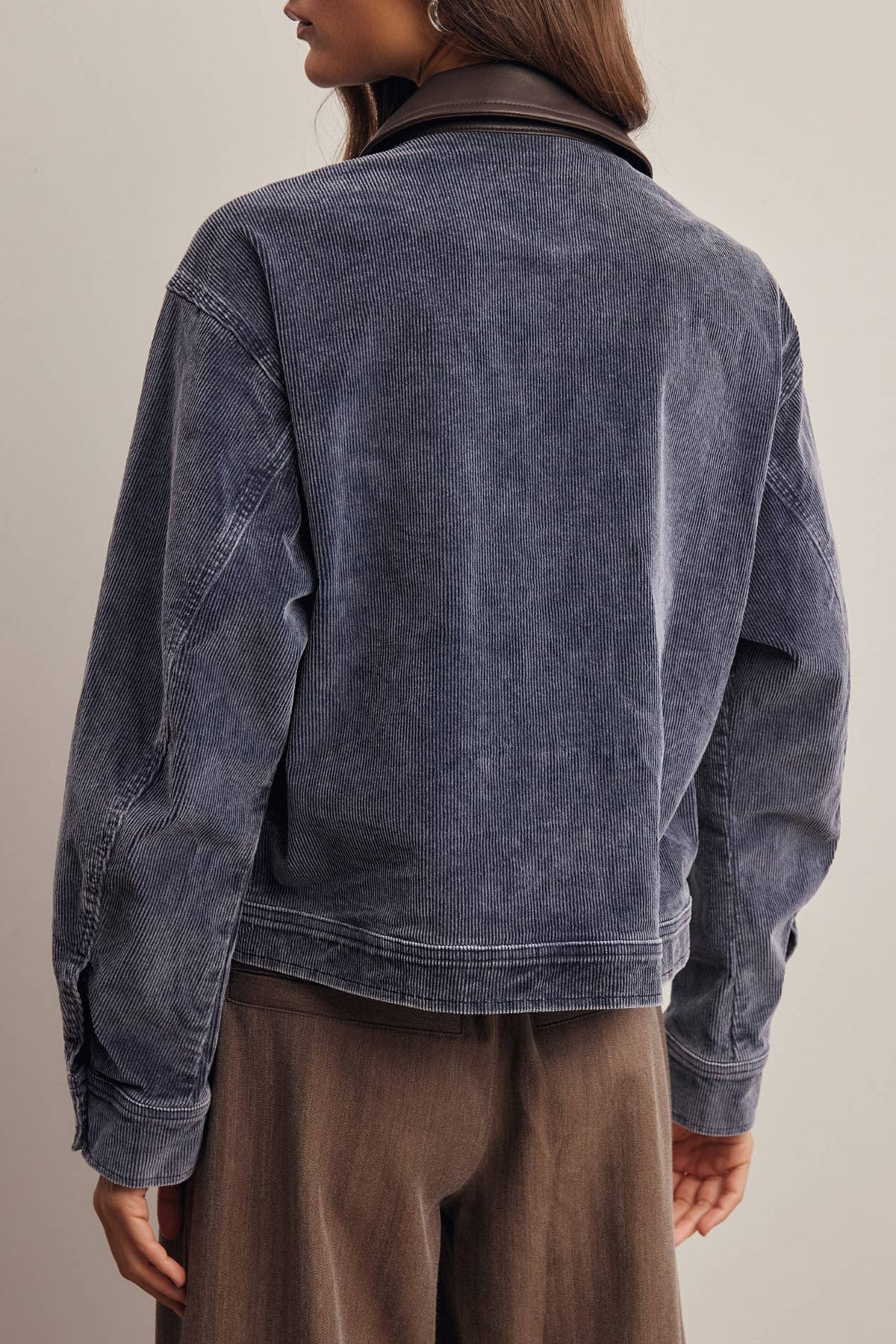 A woman shown from the back wearing a washed slate blue denim jacket with a dark brown collar, paired with brown wide-leg trousers, against a neutral beige background.