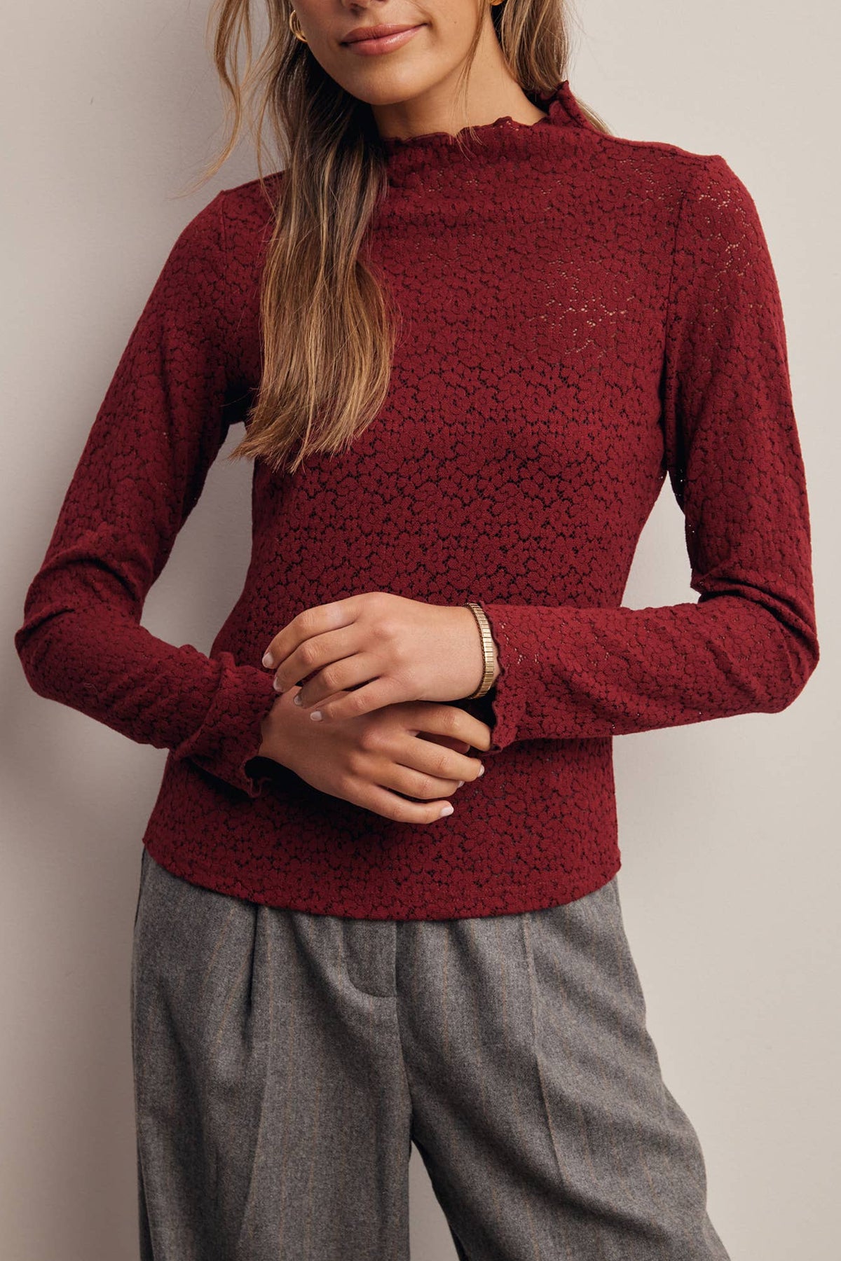 A woman wearing a textured, long-sleeve mock neck sweater in deep burgundy, paired with gray trousers, standing against a neutral light gray background.