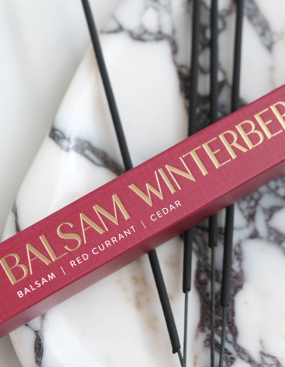 A close-up of a red rectangular product box with gold lettering reading 'BALSAM WINTERBERG' and smaller text 'BALSAM | RED CURRANT | CEDAR', placed diagonally on a white marble surface with black decorative rods behind it.