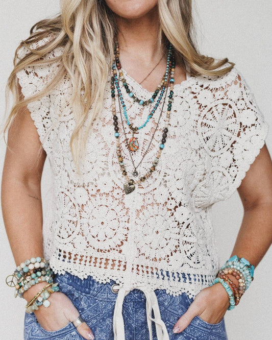 Woman wearing a white crochet top and blue patterned pants with multiple necklaces and bracelets.