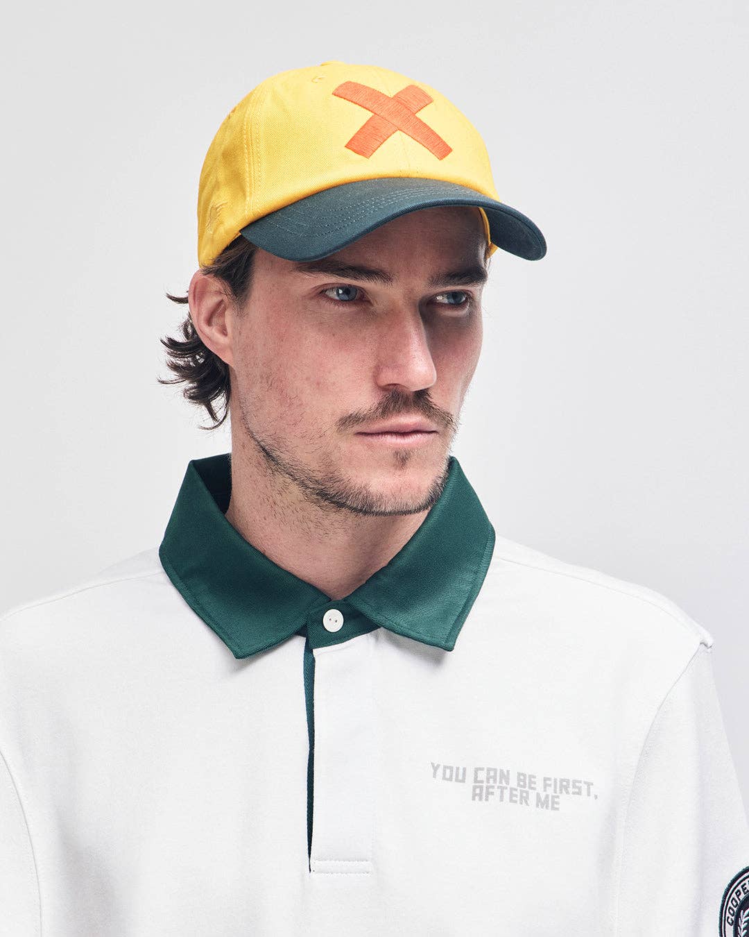 Man wearing a yellow cap with a red 'X' and a white shirt with green collar.