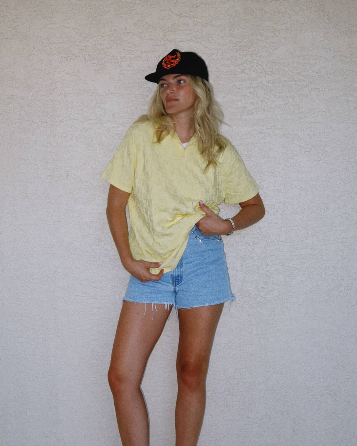 Person wearing a yellow shirt and blue shorts standing against a plain white wall.