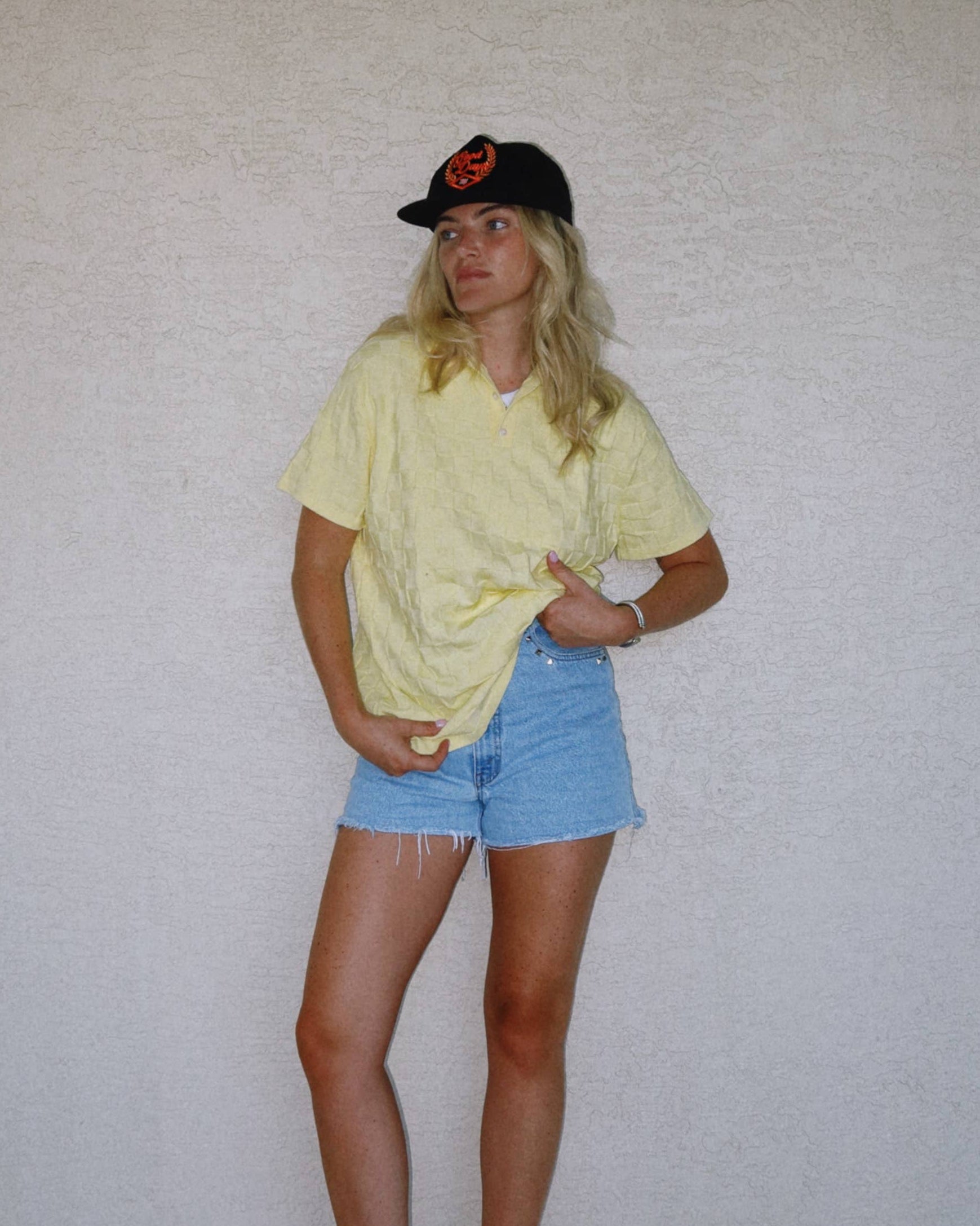Person wearing a yellow shirt and blue shorts standing against a plain white wall.