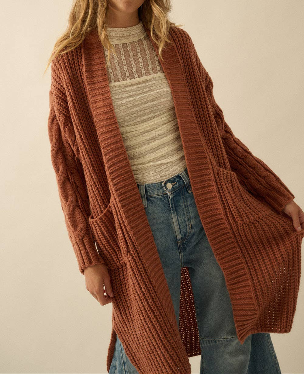 A woman wearing a long, rust-brown cable-knit cardigan over a cream ribbed top and blue jeans, standing against a plain beige studio background.