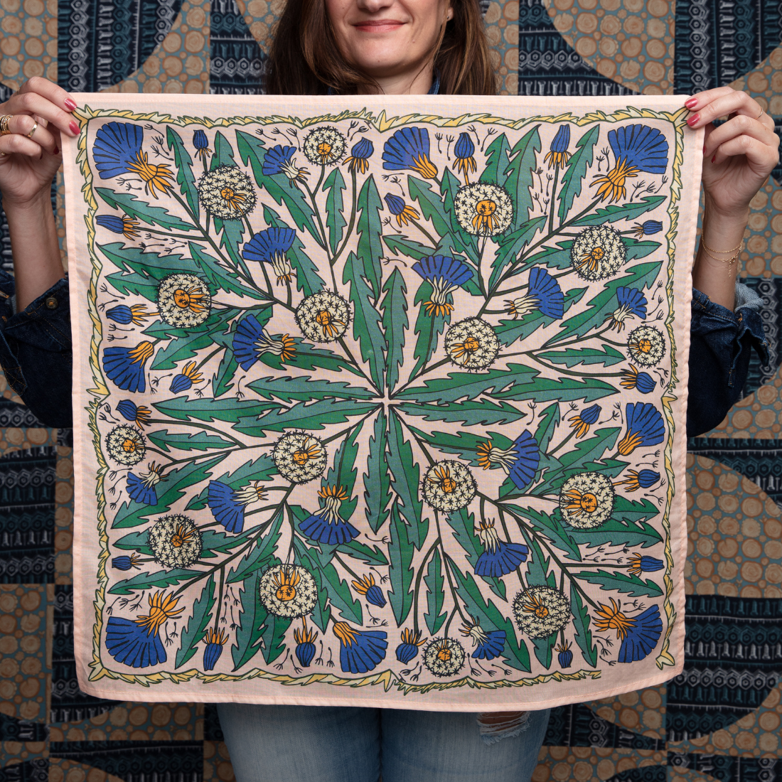 A person holds up a square scarf featuring a detailed botanical pattern with green leaves, blue flowers, and yellow accents on a light pinkish-beige background. The person is wearing a patterned top and blue jeans, with the scarf displayed flat against their torso.