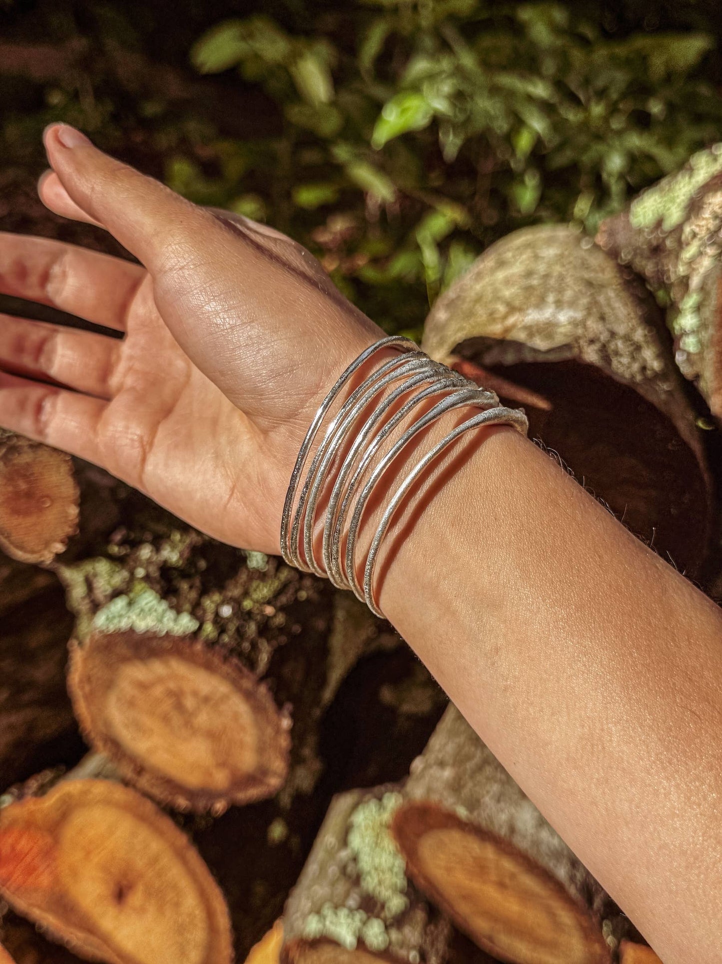 Hand wearing multiple bracelets with a natural background of logs and foliage