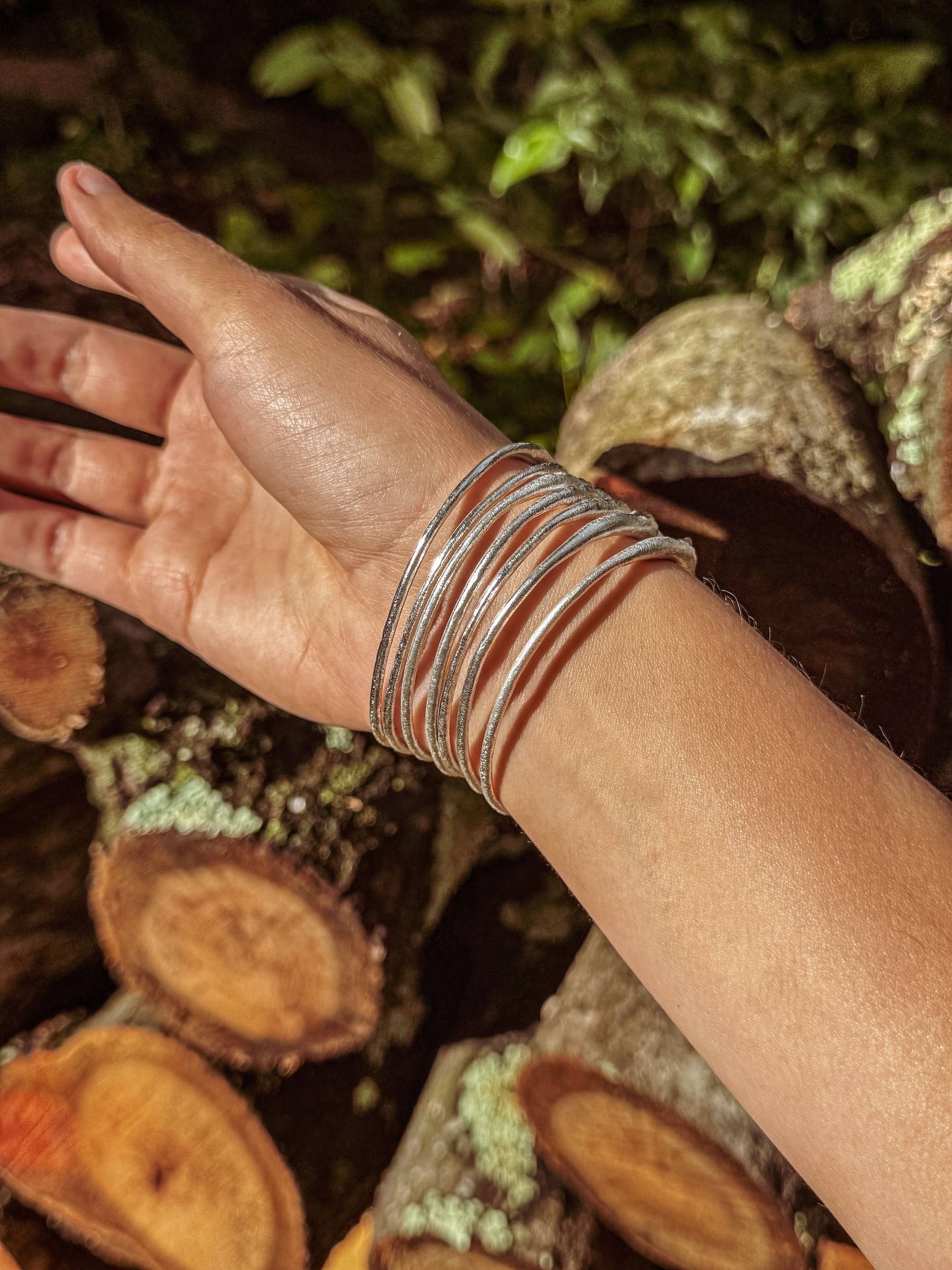 Hand wearing multiple bracelets with a natural background of logs and foliage